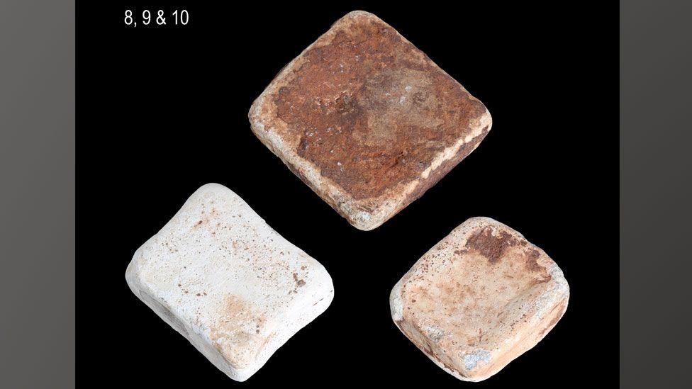 Three lead ingots against a black background. The top one is roughly square and has a bronzy patina; below on the left is a roughly rectangular ingot and is a whitish shade; below on the right a slightly smaller squarer ingot mottled cream coloured.