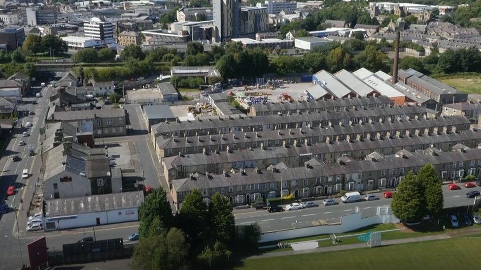 An aerial view of Burnley showing rows of terraced houses to the foreground behind a road. In the background are office blocks and commercial buildings