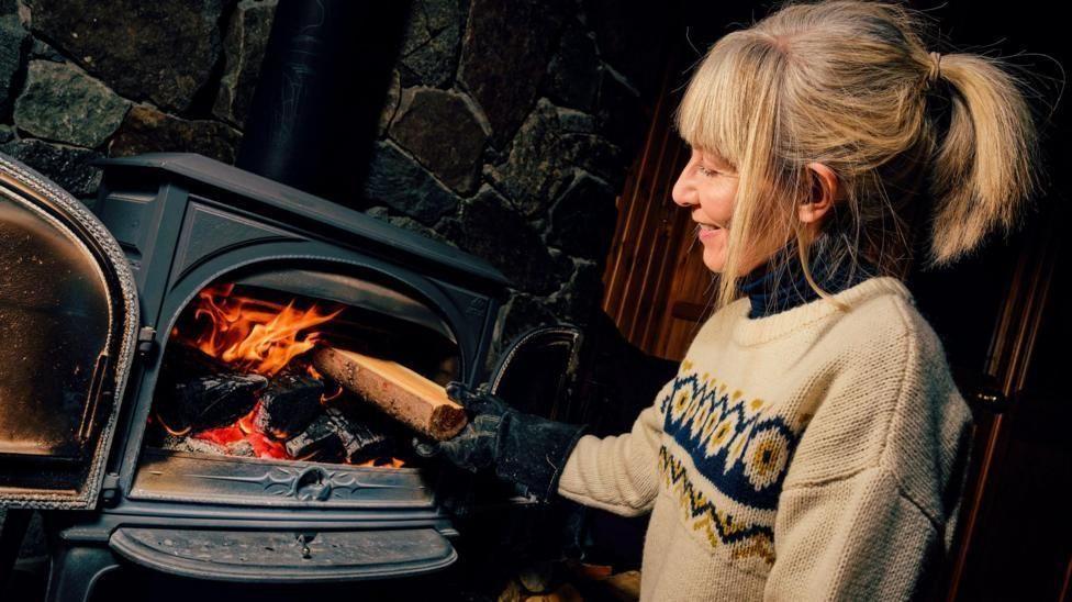 a woman wearing a winter jumper and a thick glove places a log inside a wood burning stove, which is ablaze