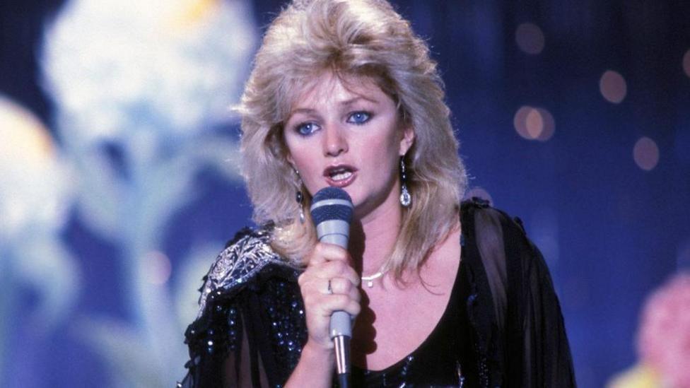 Tyler with big, blonde hair, wearing a black sparkly dress and singing into a microphone