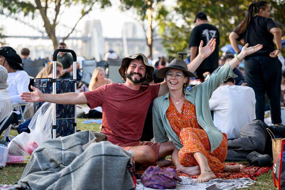 Revellers gather at Mrs. Macquarie's Point ahead of the New Year's Eve celebrations in Sydney, Australia, 31 December 2024. A man in a brown t-shirt and a woman in an orange dress and grey shirt, both wearing hats, sit on the ground in a crown and wave to the camera
