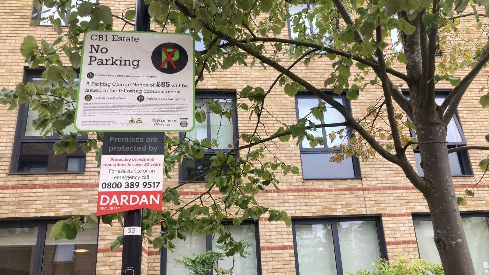 A residential estate area in Cambridge. The area is patrolled by security from Dardan. In front of a building is a tree and a sign post. On the sign it tells people there is no parking on the CB1 estate and that the premises are protected by Dardan.