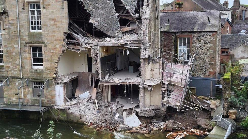 The Old Courthouse after its collapse. The rear section of the building has fallen apart and is scattered into the River Cocker. The inside of the buildings, which have bare brick walls can be seen.