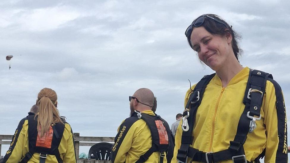Three people outdoors are wearing bright yellow jumpsuits with black straps around them. Two, on the left, are watching a parachutist in the sky. Belinda Taylor, on the right, is looking at the camera with a small smile and her head tilted slightly to one side. She has brown hair and is wearing goggles on her head