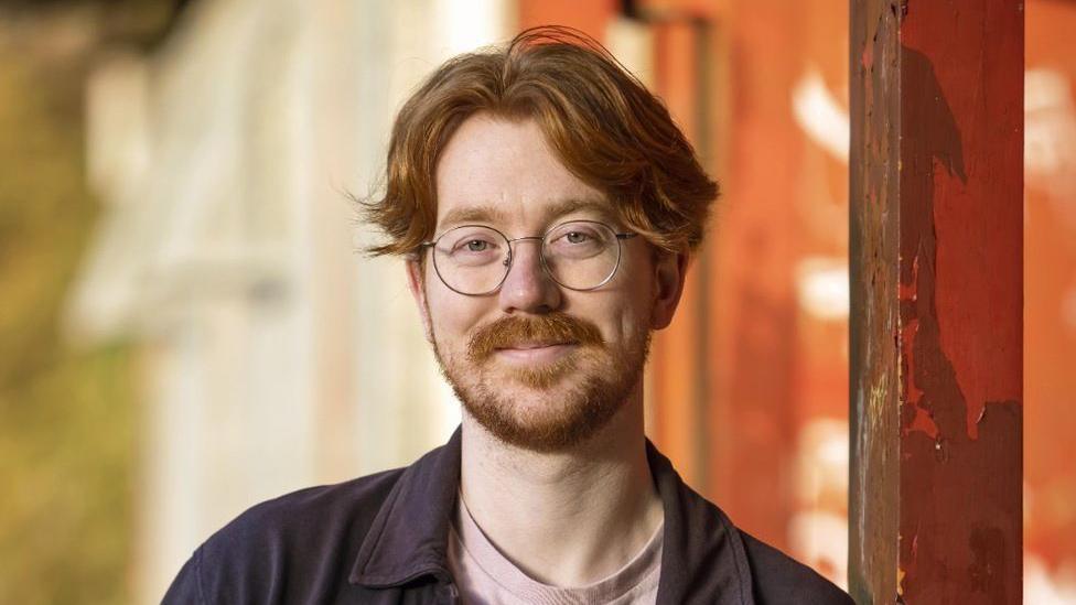 Matt McCallum smiles at the camera. He has ginger hair and is wearing silver glasses and a light pink T-shirt with a navy shirt on top.