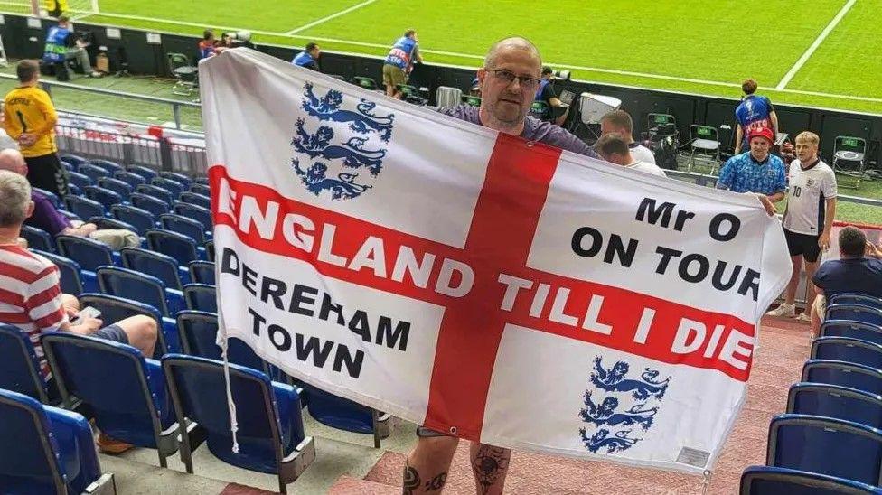 Ian Odgers stands in a stadium holding an England flag that says 'England till I die' across the center.