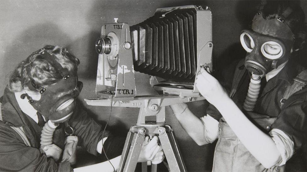 Two Women's Auxiliary Air Force (WAAF) trainees practising photography whilst wearing gas masks to simulate working under active service conditions. The photograph was taken during training at No.2 School of Photography in Blackpool