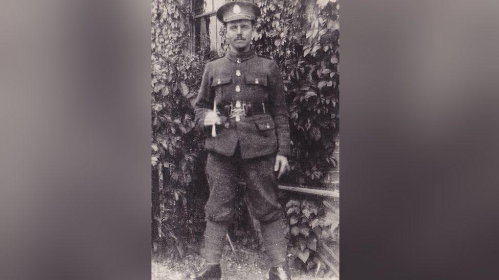 A soldier is seen in an old black and white, grainy photo. He is wearing an Army uniform typical of World War One. He has a moustache