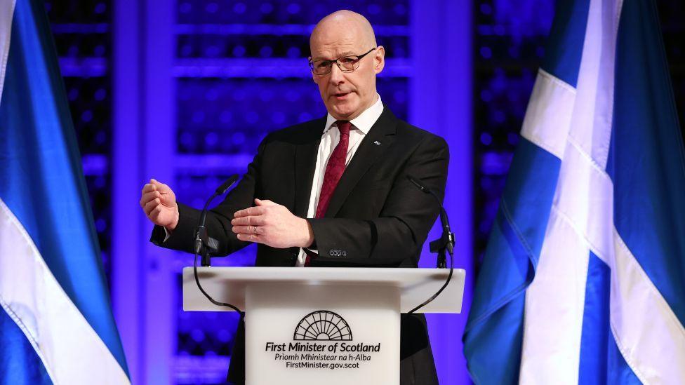 A bald man with glasses, wearing a dark suit and pink tie, stands with his hands raised in front of his body as he speaks at a podium. Saltires are illuminated in the background.