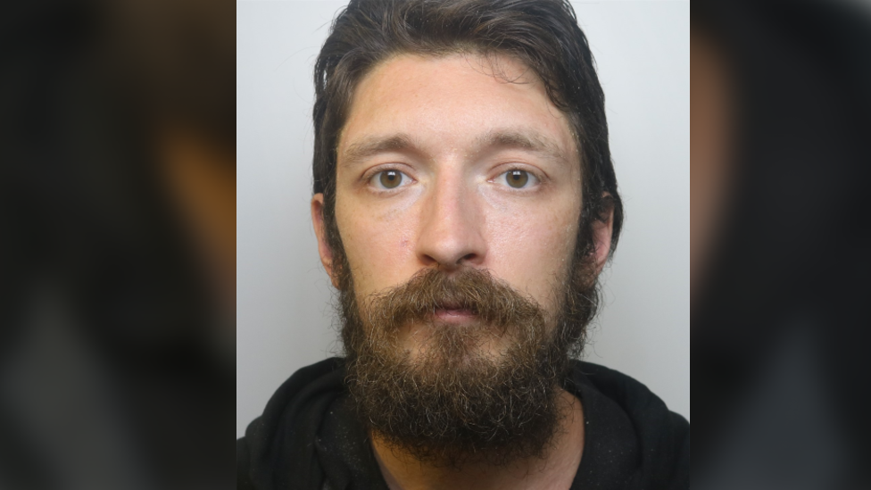 A police custody photo of David Parish. He has short dark brown hair and a thick dark brown beard. He is wearing a black hooded jumper.