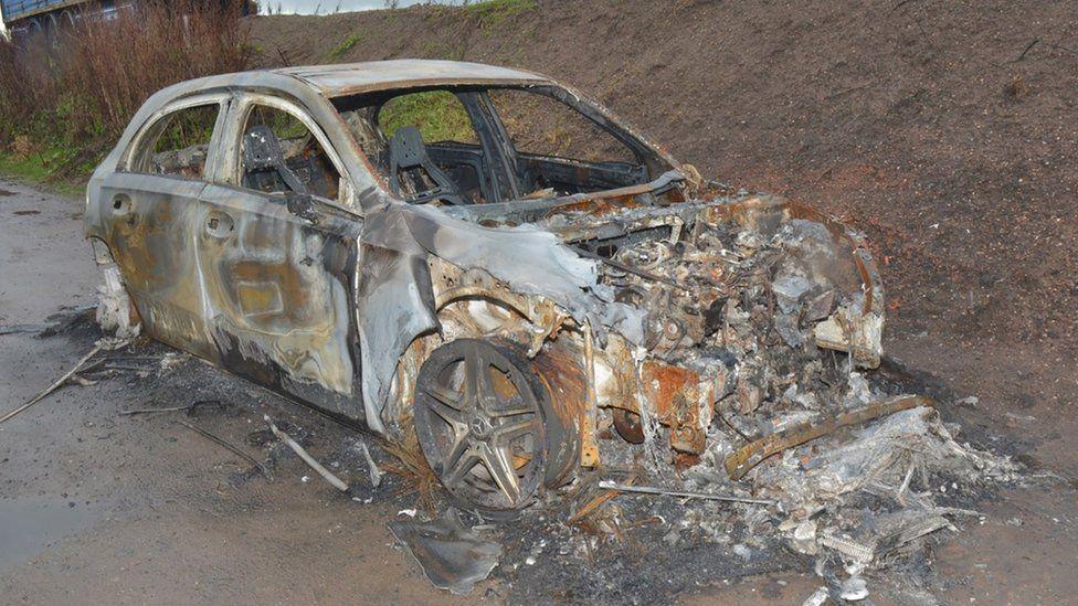 The charred and twisted remains of a hatchback Mercedes car parked next to an embankment. The windows are gone and the black paint has been burnt away making it appear grey in colour