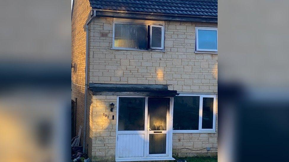 A two-storey stone house on a housing estate which has white windows and a porch with a white UPVC door. There is smoke damage and soot over the front door and an upstairs window above.