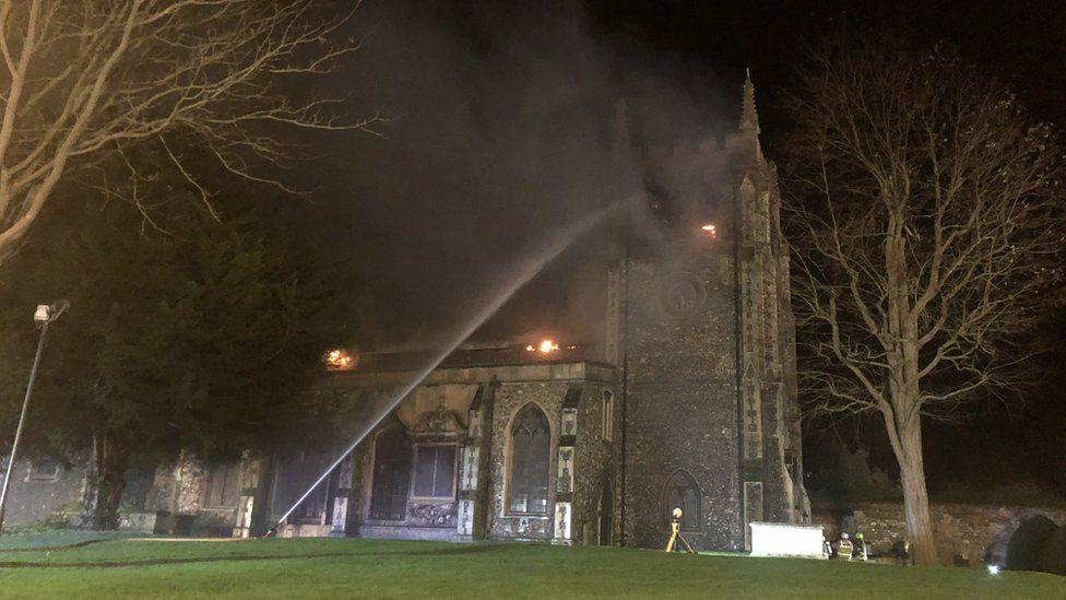 Firefighters spray a hose at at St John the Baptist Church, Royston, while flames flicker on the roof at night.