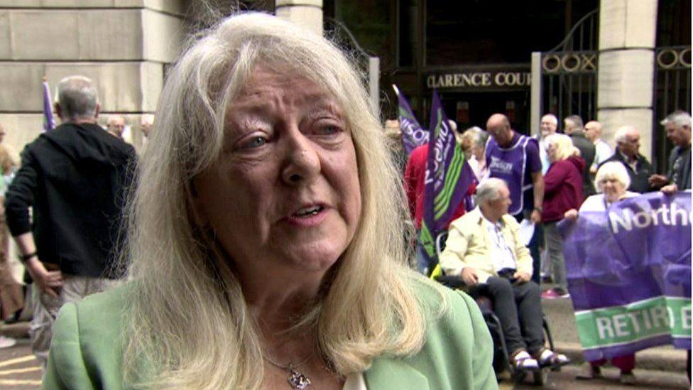 Patricia McKeown with long grey hair in green jacket in front of a picket line