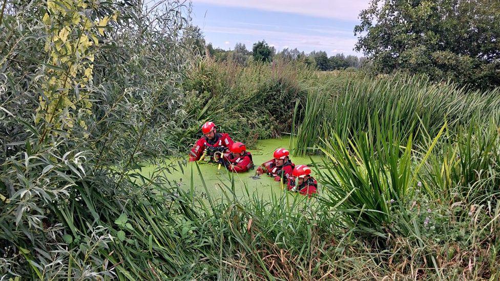 Four police divers in red clothing and hard hats walking in chest height murky water, surrounded by undergrowth.