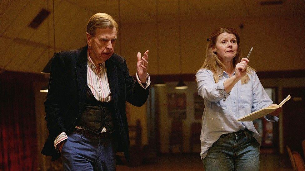 Timothy Spall and actor Sian Gibson standing up on Death Valley set. Her character is holding a book with a pen upraised in the other hand and is making conducting motions, which he appears to be mirroring.