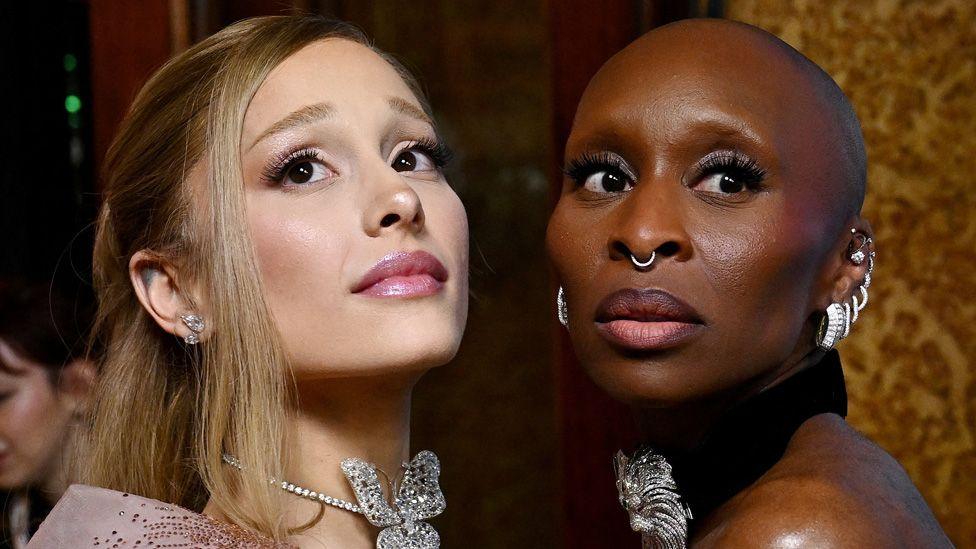 Ariana Grande and Cynthia Erivo facing each other and both looking sideways in the same direction, wearing jewelled necklaces and earrings at the Australian premiere of Wicked in November.