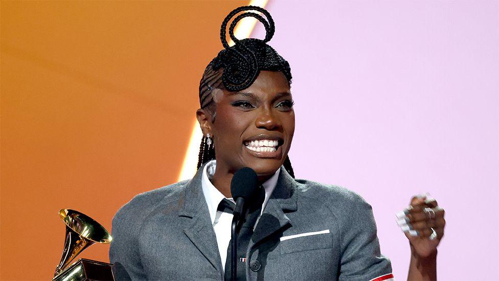 Doechii on stage after winning a Grammy award, with a trophy in her right hand, as she speaks into a black mic. She is smiling and wearing a grey blazer and white shirt and black tie.