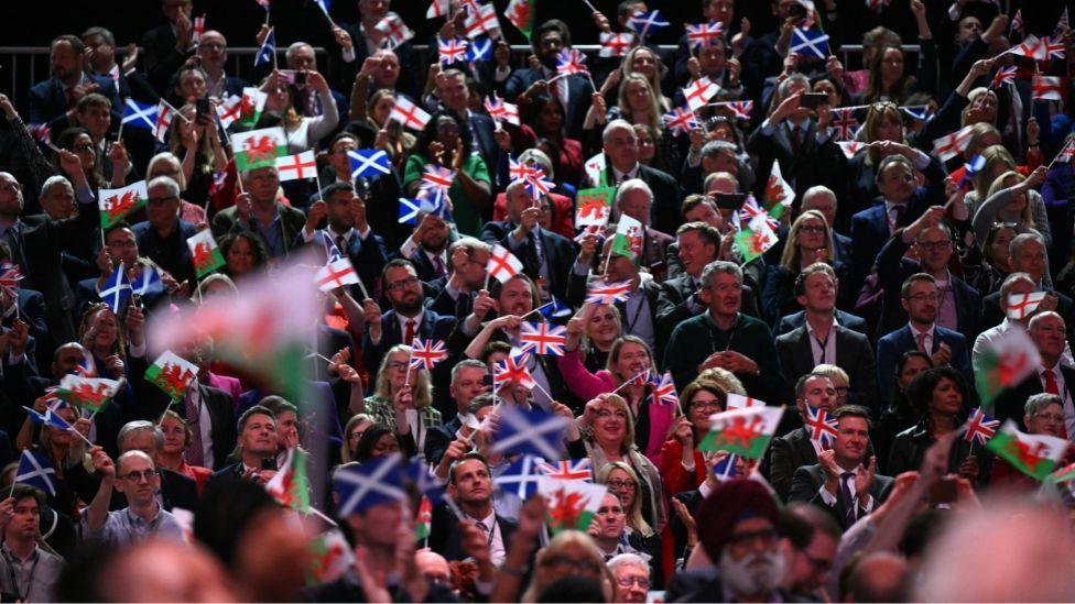 People tightly packed into a stand wave small flags above their heads. There are blue and white saltires, Welsh red dragon flags and red and white St George's cross flags.