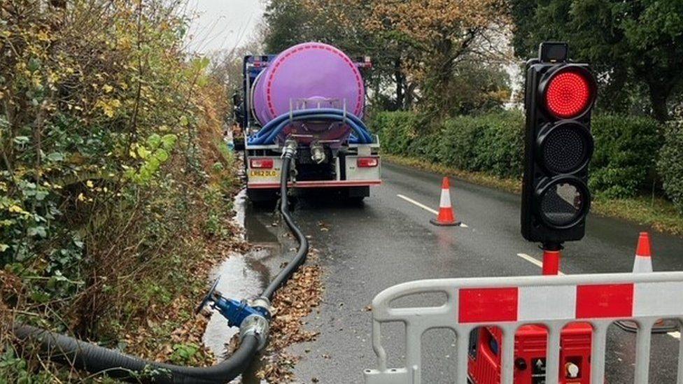 Barrier across road with traffic light behing and purple tanker with hose stretching from rear into hedge.