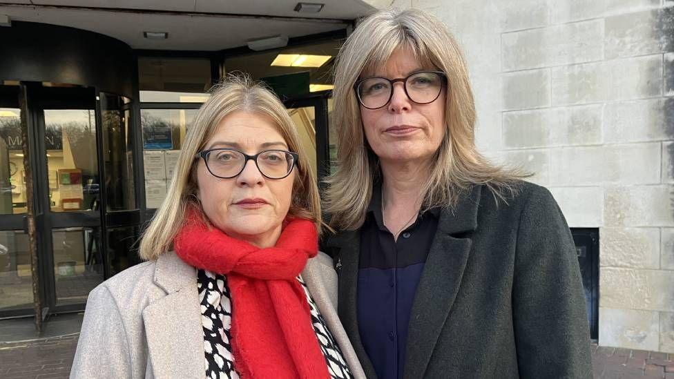 Two women, both with short blonde hair and wearing glasses, looking at the camera. Fiona, on the left, is wearing a red scarf, off-white jacket and black and white blouse; on the right, Liz is wearing a grey jacket and blue and black shirt 