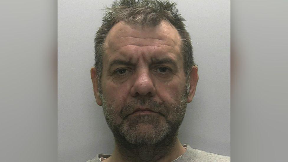 A close-up of Lee Blatchford who has a brown and grey short beard and brown and grey hair. He is directly into the camera and is not smiling.