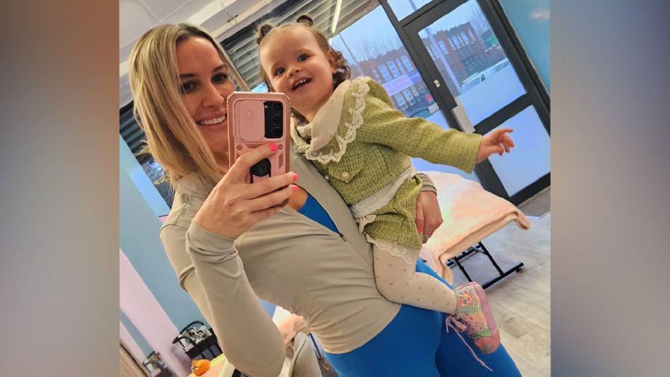 Natasha has long blonde hair and is wearing blue lycra leggings, a blue top and a beige lycra jacket. She is holding Sienna-Ro,se who is smiling at the camera and wearing a green tweed outfit.