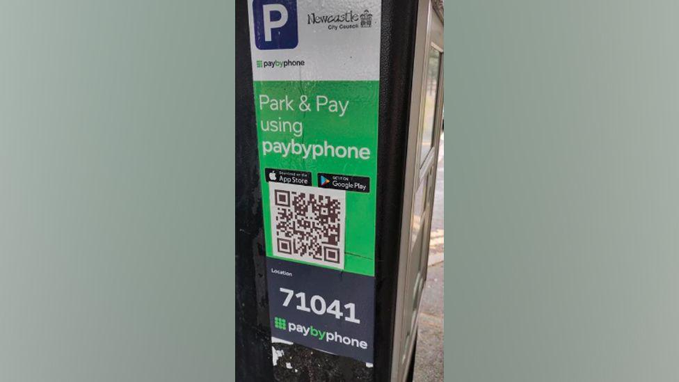 A sticker placed on a parking meter showing a QR code. The sticker uses official logos from Newcastle City Council and the parking ticket company PaybyPhone. It also includes logos for the Apple App Store and Google Play.