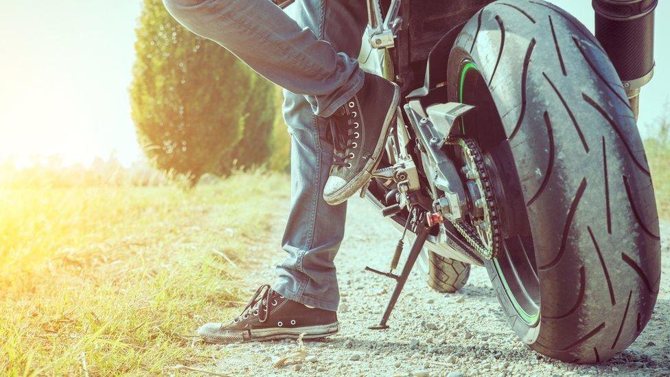 A biker's feet next to the wheels of a motorbike while it stands on its kickstand. The feet wear converse trainers with white toe tips and the rest of the footwear is dark. They stand on a gravel path next to a grassy field. The bike's wheels have dust on them.