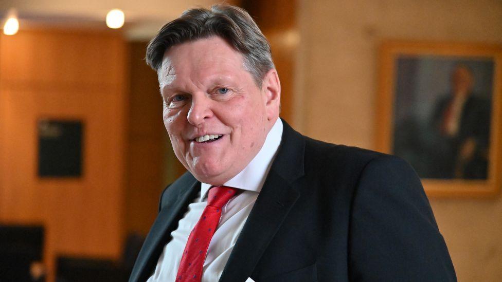 Stephen Kerr, who has dark hair with a side parting, looks at the camera in a medium close-up shot in the Scottish Parliament. He is wearing a black suit, white shirt and red tie. 