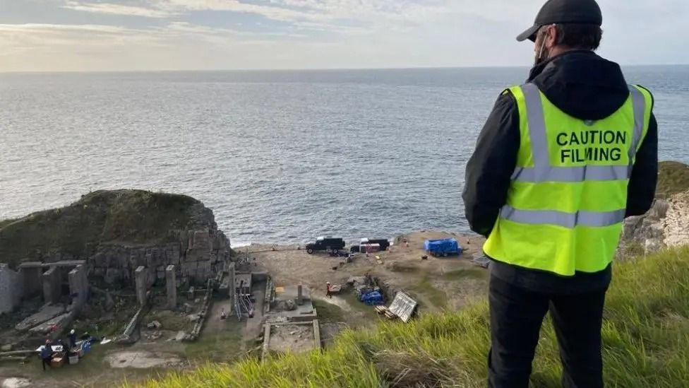 A security guard with 'caution filming' on the back of his hi-vis vest looks down at a disused quarry, with vans and film staff working among the rocks