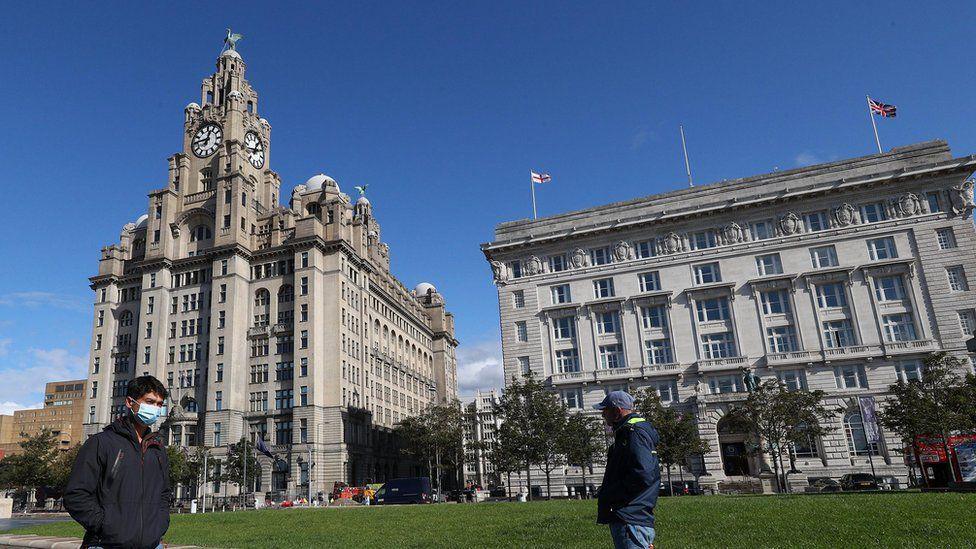 Two men walk in opposite directions past the Royal Liver Building, a grey stone box shaped structure with two clock towers, each crowned with a statue of the famous Liver Birds. 