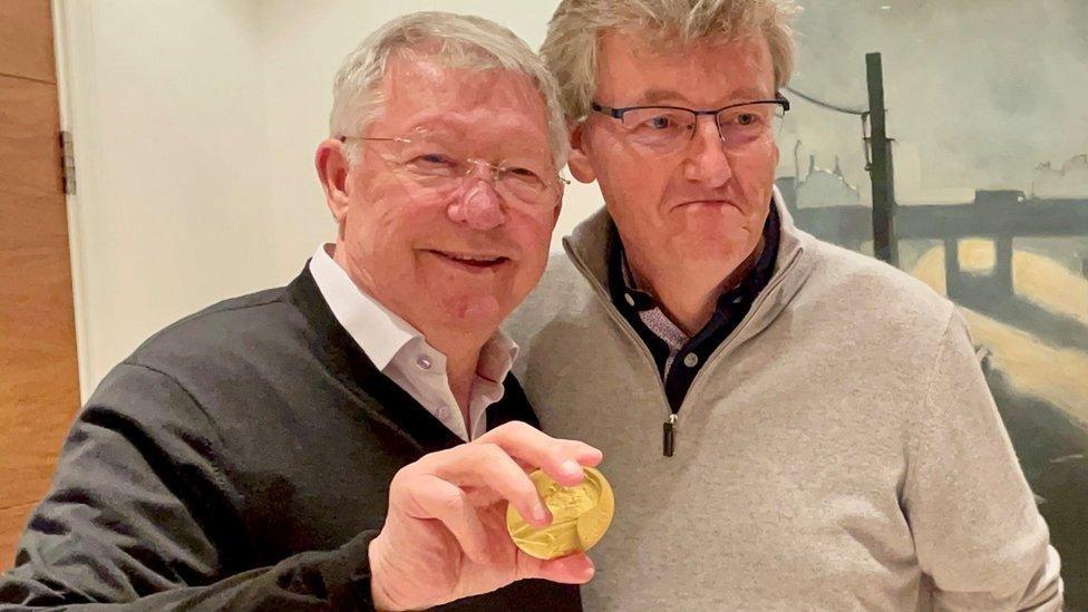 David MacMillan and Sir Alex Ferguson stand side by side. Both are wearing glasses. Sir Alex wears a black jumper and shirt. He is on the left of the image holding David's gold Nobel Prize Medal and smiling. With David on the right wearing grey. 