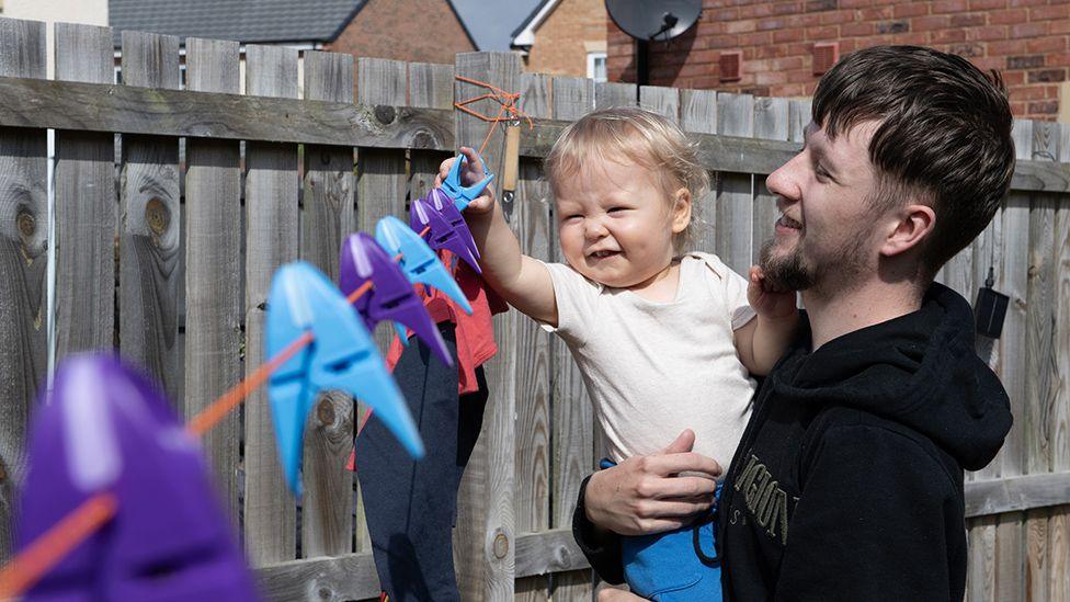 Callum Fay is holding his son Archie in his arms as he plays with pegs on a washing line. They're both smiling.