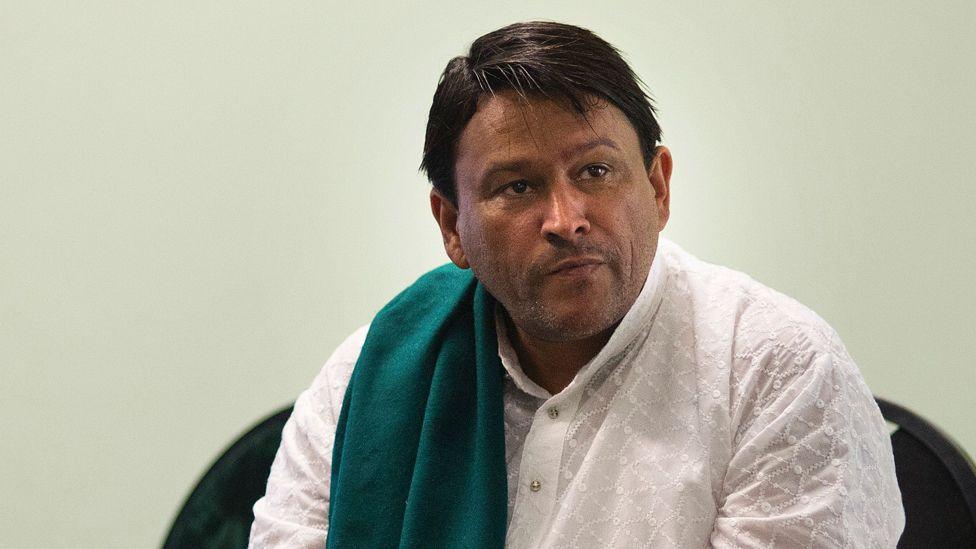 A close-up of Imam Muhsin Hendricks wearing a white shirt and a green scarf draped over one shoulder. The shot was taken in 2016.