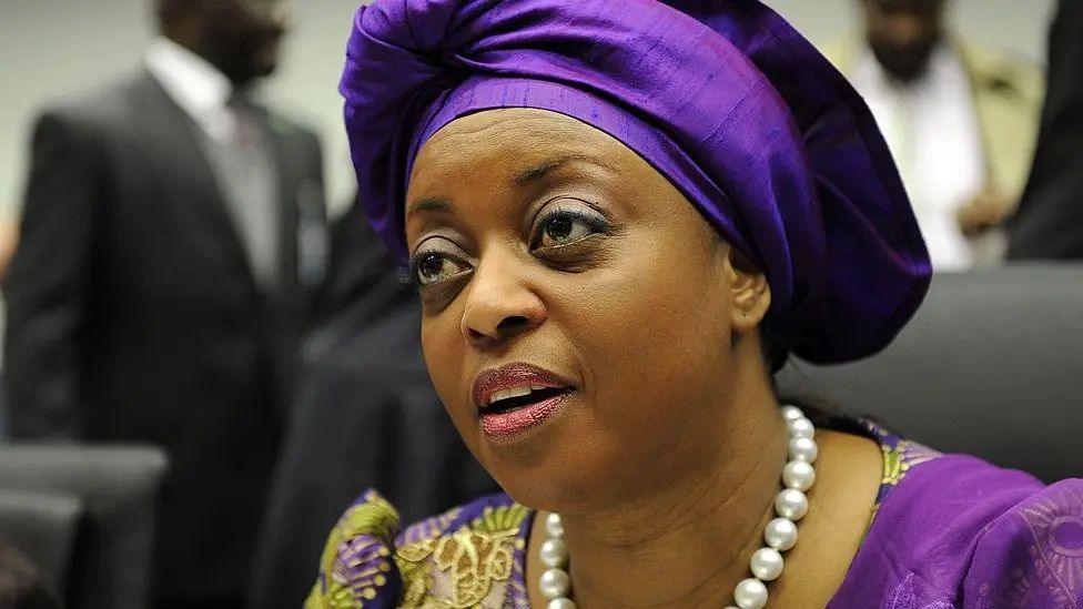 A woman dressed in a purple outfit with a matching gele (headdress) sits on a chair and speaks.