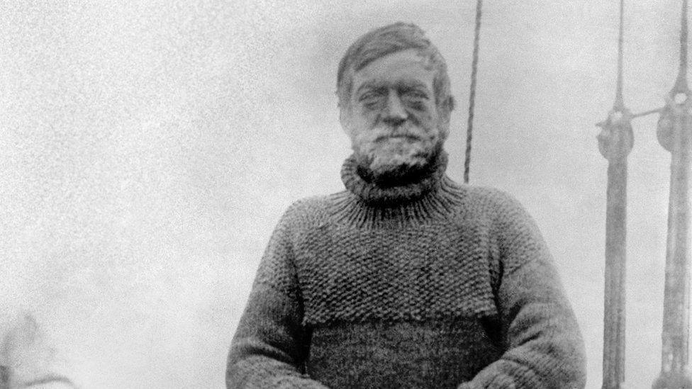 A vintage black and white photograph of a man in his late forties on a ship. The man has a weather-beaten face and a beard and is wearing a thick woollen jumper.