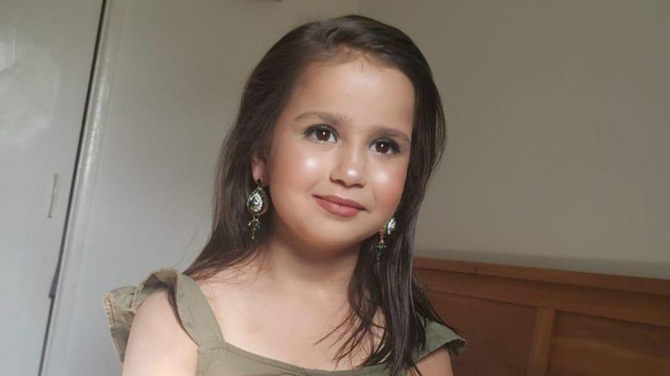 Sara Sharif is smiling. She is long hair below her shoulders. She is wearing green and has large dangly earrings and seems to be wearing make-up.