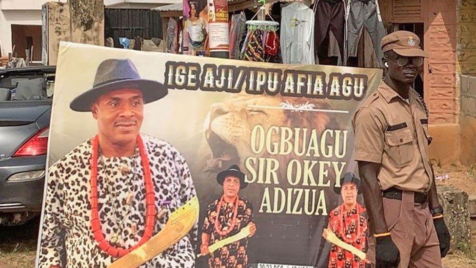 A uniformed security officer stands to the right of a poster advertising a ceremony for a man called Sir Okey Adizua to become a 'Leopard Slayer'. There are three images of a man holding a gold sword, wearing a large red bead necklace and a wide-brimmed black hat