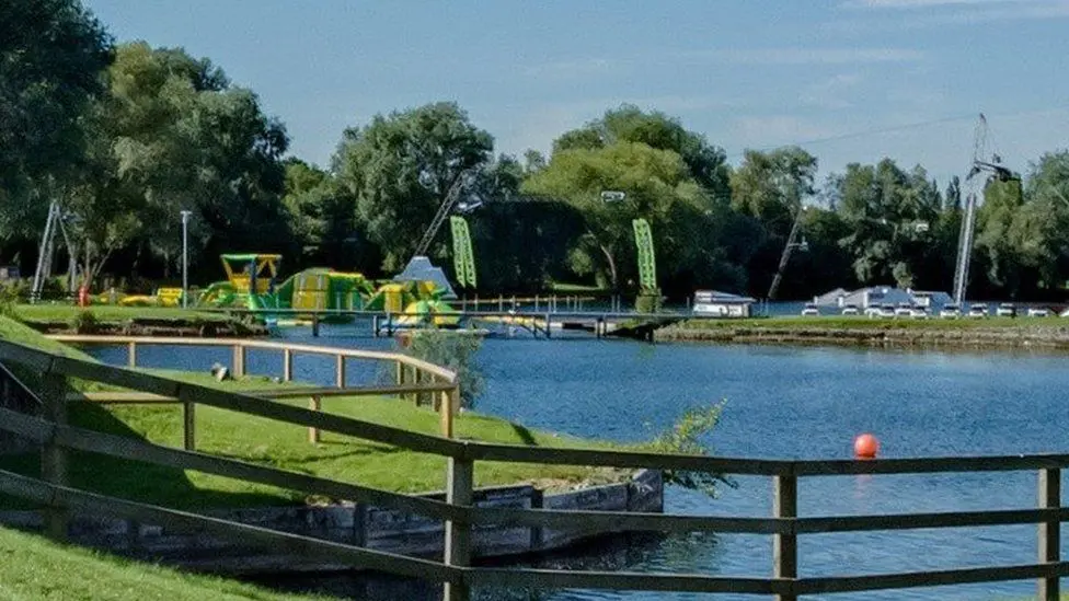 The picture shows Liquid Leisure watersports centre. There is a body of water with grass visible on either side. In the background are green and yellow obstacle courses floating on the water.