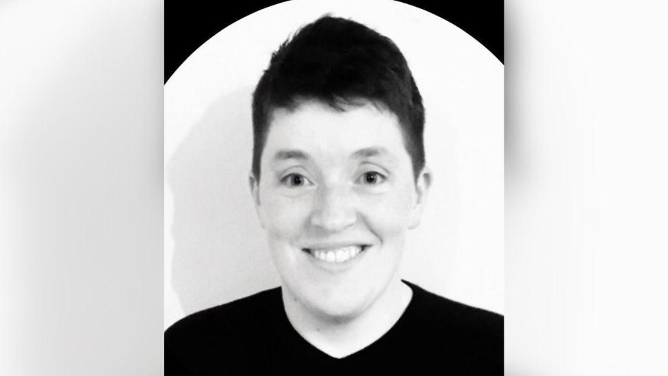 Rev Teagan MacAodhagáin in a black and white image, he has short dark hair and wearing a black t-shirt 