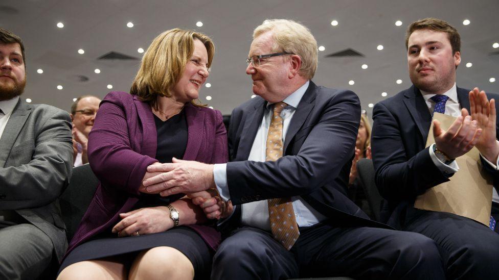 Michelle Ballantyne, with light brown highlighted hair, and Jackson Carlaw, with greying blond hair, shake hands while seated. They are photographed from a low angle while surrounded by other seated people. Ballantyne is wearing a purple jacket and dark dress, while Carlaw is wearing a dark suit and gold tie. 