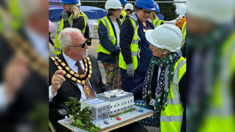 Councillor Stephen Cooper wearing a black suit, black sunglasses and his mayoral chains. He is sitting down with a model replica of the treatment centre resting on his lap. He is surrounded by women wearing hard hats and yellow hi-vis vests on a construction site.