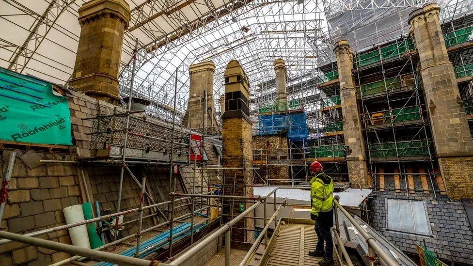 A workman stands on a platform and looks out over scaffold-covered rooftops at Manchester Town Hall. 