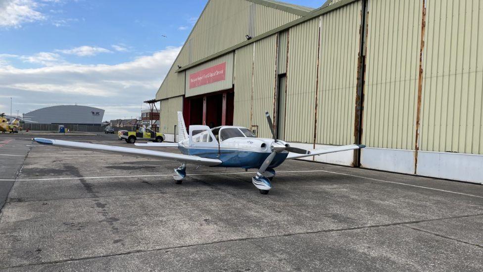 A small blue and white plane in front of a large beige hangar at Blackpool Airport