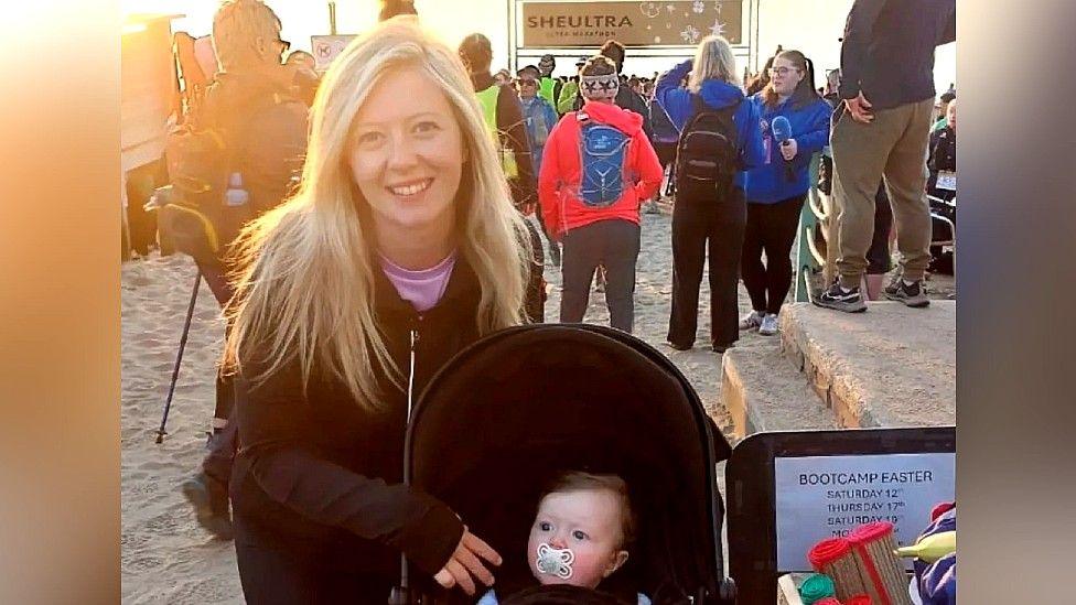 Enlli is on the beach with Cali in a black pram. Enlli has her hair loose and is smiling directly at the camera. She wears a dark, zip-up jacket over her purple runner's top. The rising sun is slightly bleaching out the top left of the photo. There are lots of people in the background, some with running equipment and bags. A starting banner with the words She Ultra on it can be seen in the near distance.