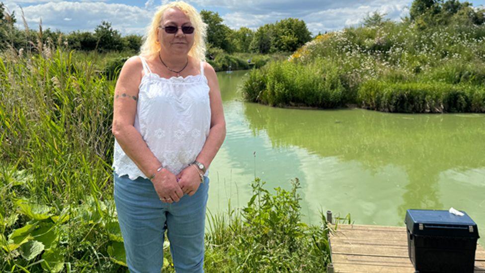 A woman wearing sunglasses, a white strappy top and blue jeans. She is standing by a fishing lake with a small wooden jetty and a black box visible to the right