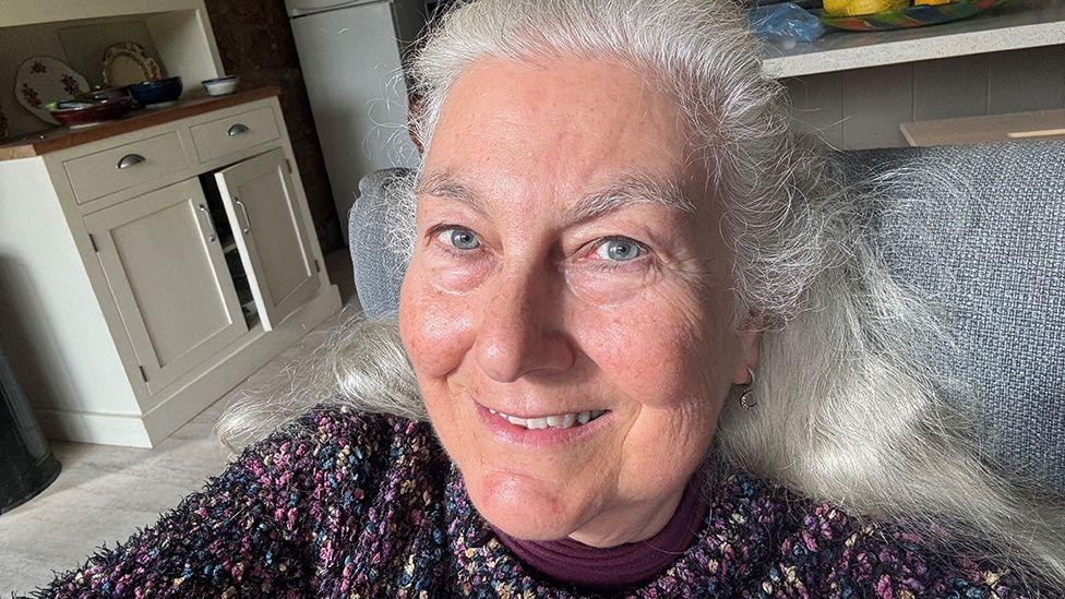 Elfie Waren, who has long grey hair and is wearing a purple mottled knitted jumper, smiling in a selfie taken in her kitchen, with cream wooden units in the background.