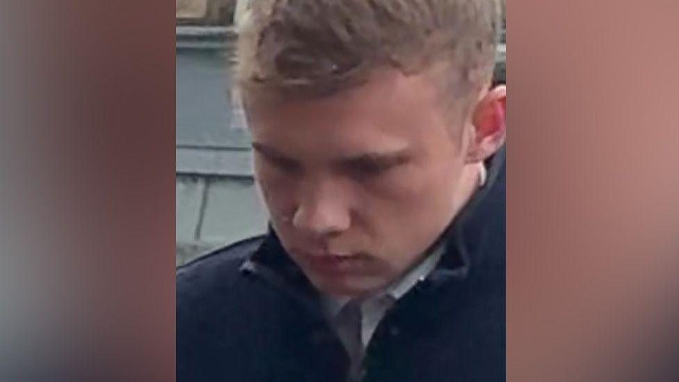 A man, Charles Gray wearing a dark jacket and light-coloured collared shirt, standing outdoors near a stone wall or building. He has a glum expression with his head lowered.