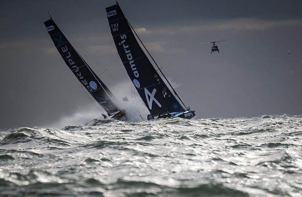 Two sleek racing sailboats with tall black sails lean sharply as they cut through choppy gray waves under a dark, cloudy sky. Sponsor names are visible on the sails. A helicopter hovers above, adding to the dramatic offshore scene.